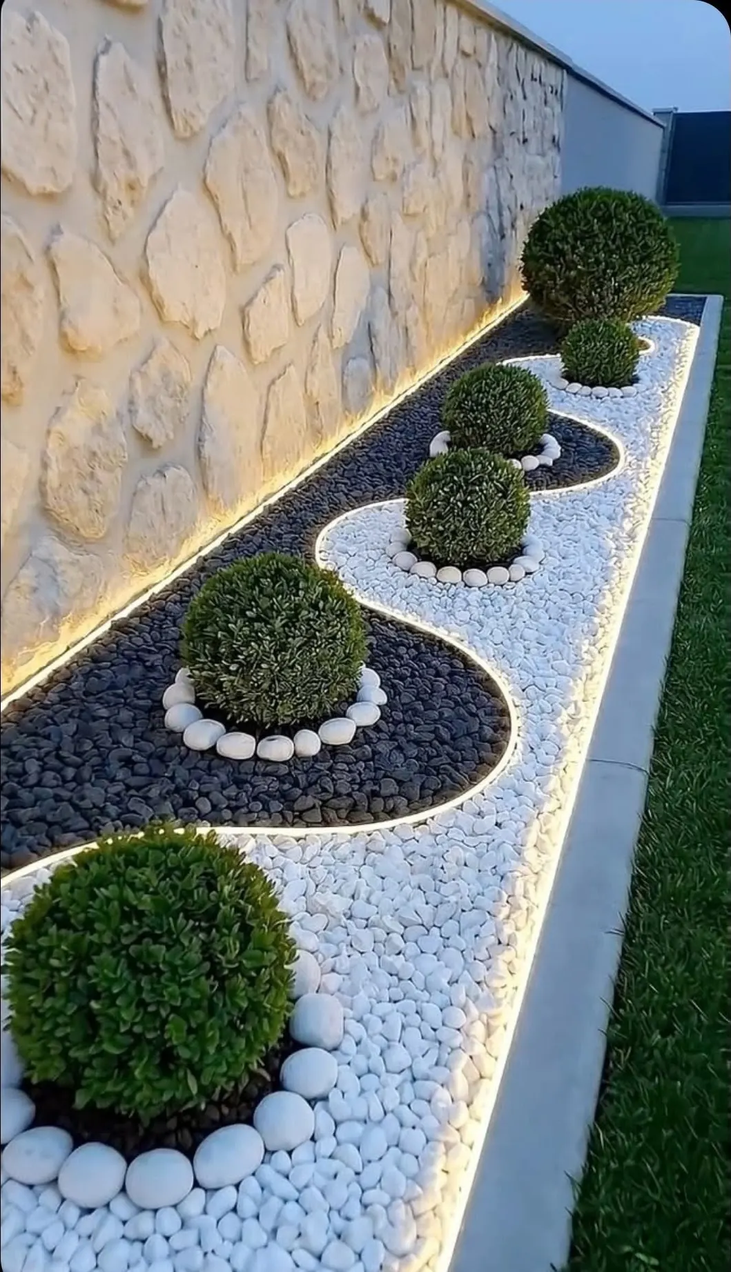 Lit pebble bed with topiary boxwoods set against a stacked-stone wall